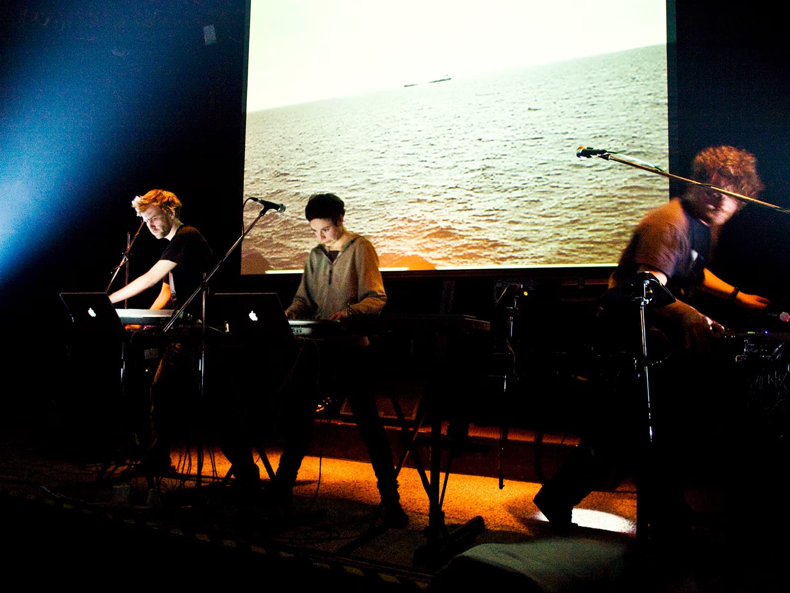Trzech muzyków wykonuje muzykę elektroniczną na ciemnej scenie z projekcją oceanu w tle.