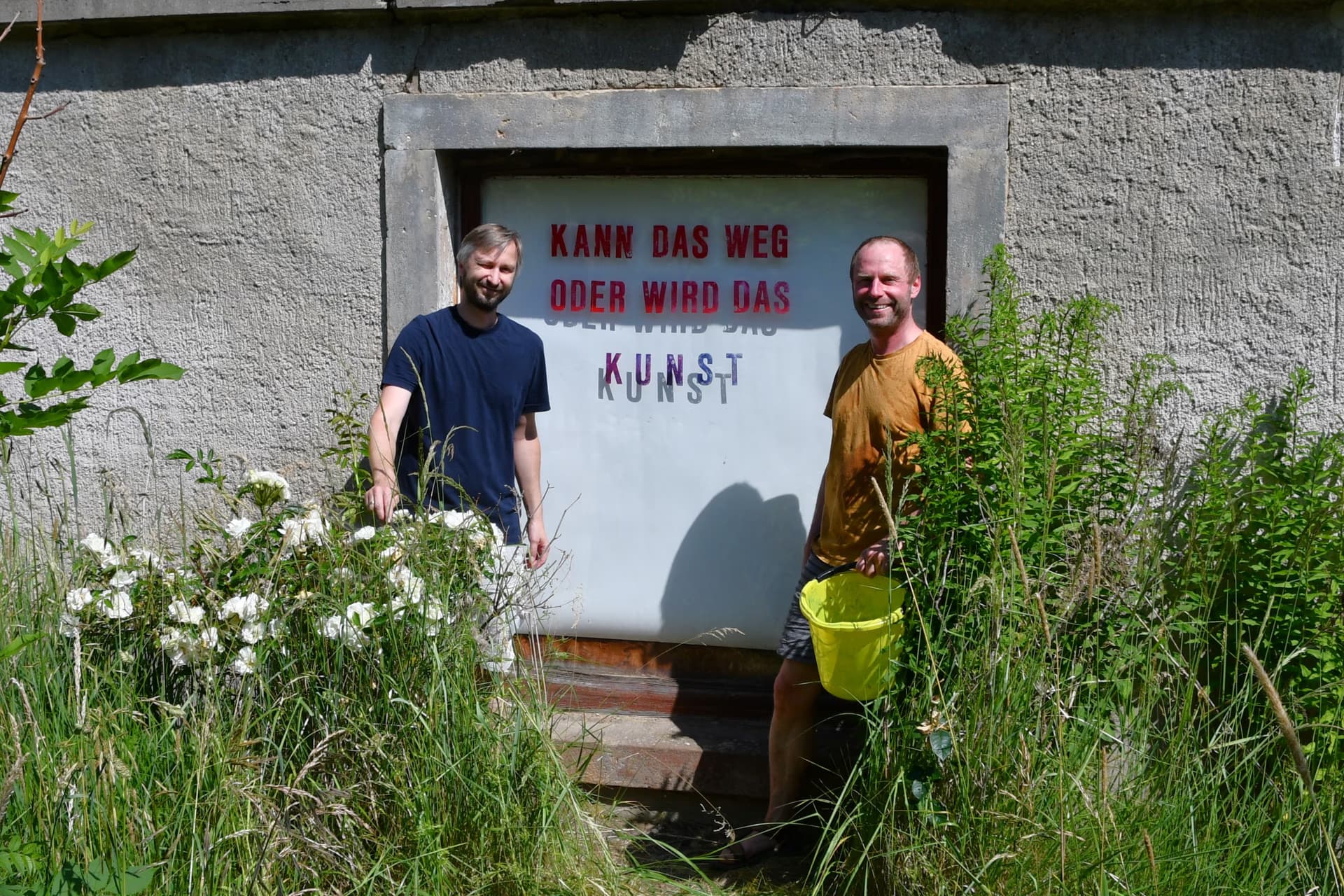Zwei Männer stehen vor einem Kellereingang mit der Aufschrift Kann das weg oder wird das Kunst.