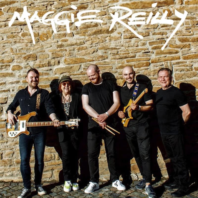 Five band members pose against a rustic stone wall with their musical instruments.