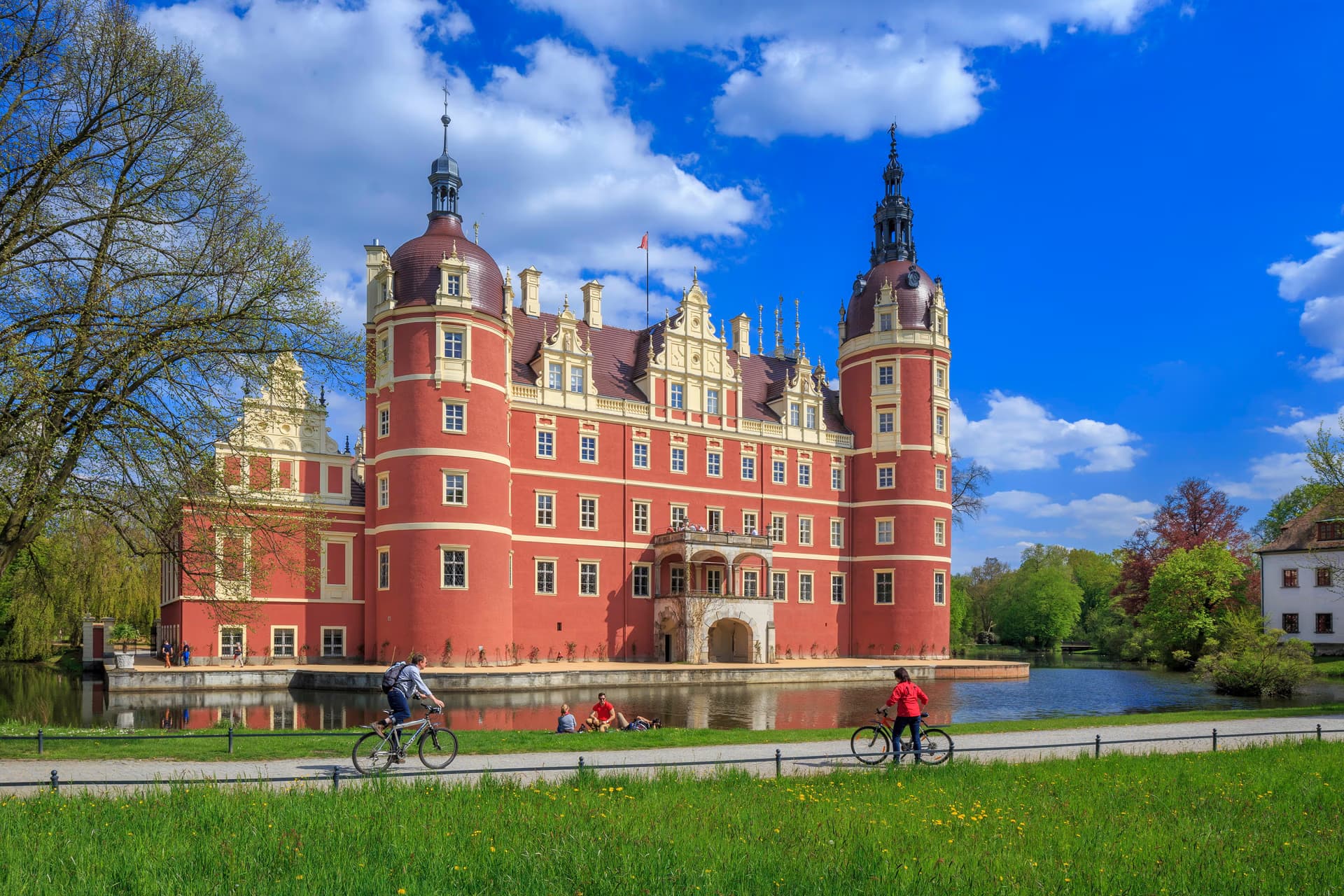 Zamek w Mużakowie wznosi się nad fosą, a obok na ścieżce rowerowej jadą rowerzyści.