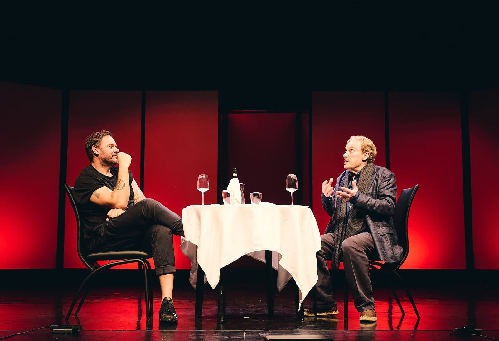 Two men talk while sitting at a small table on a stage with a red background.