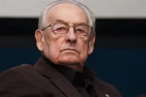 Elderly man with white hair and glasses wearing a dark jacket, looking forward seriously.