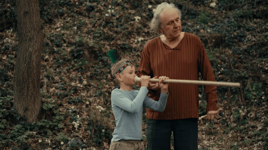 A young boy plays a long wooden pipe while an older man watches in a forest setting.