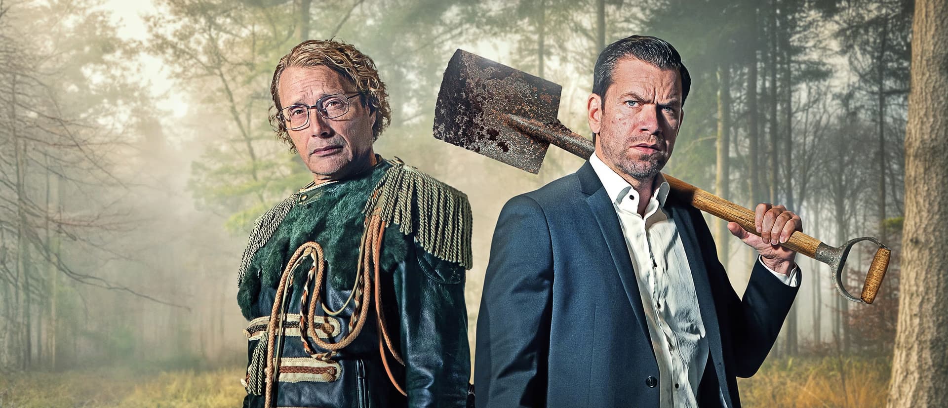 Zwei Männer stehen im Wald, einer trägt eine Jacke mit Fransen, der andere einen Anzug mit einem Spaten.