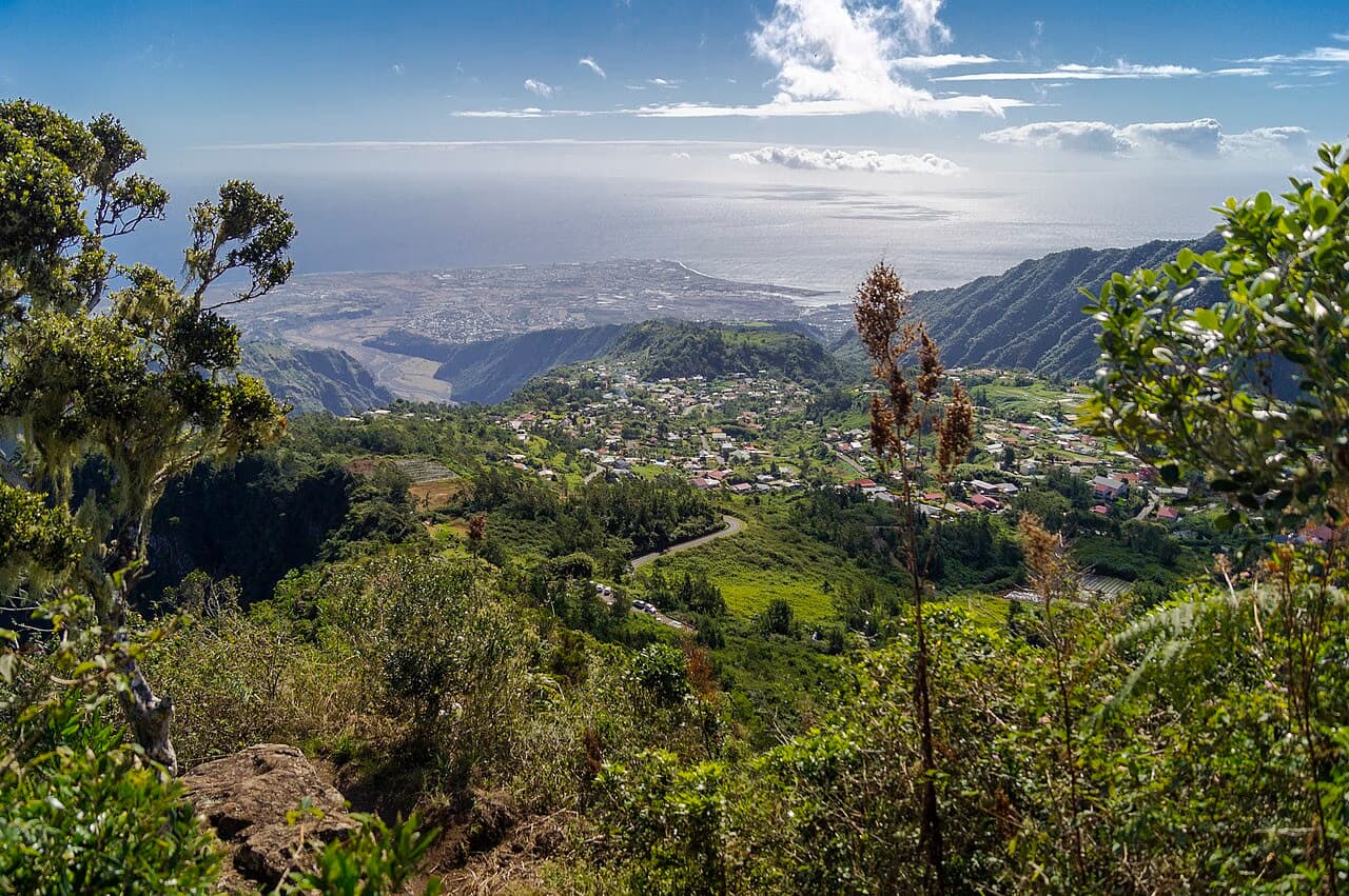 A lush mountain valley with a small village overlooks the distant ocean under a blue sky.