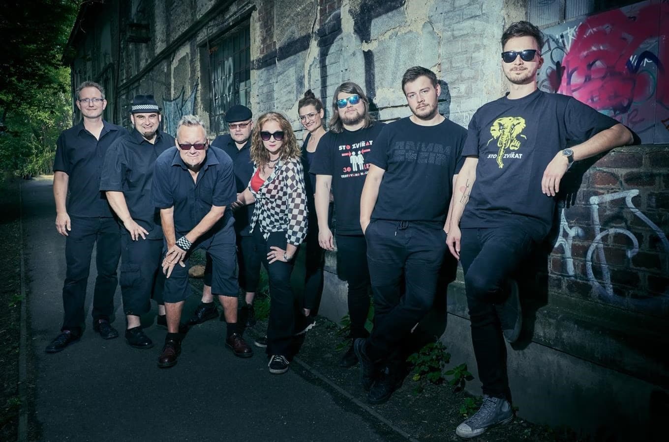A large band group posing together standing in front of a graffiti-covered brick wall.