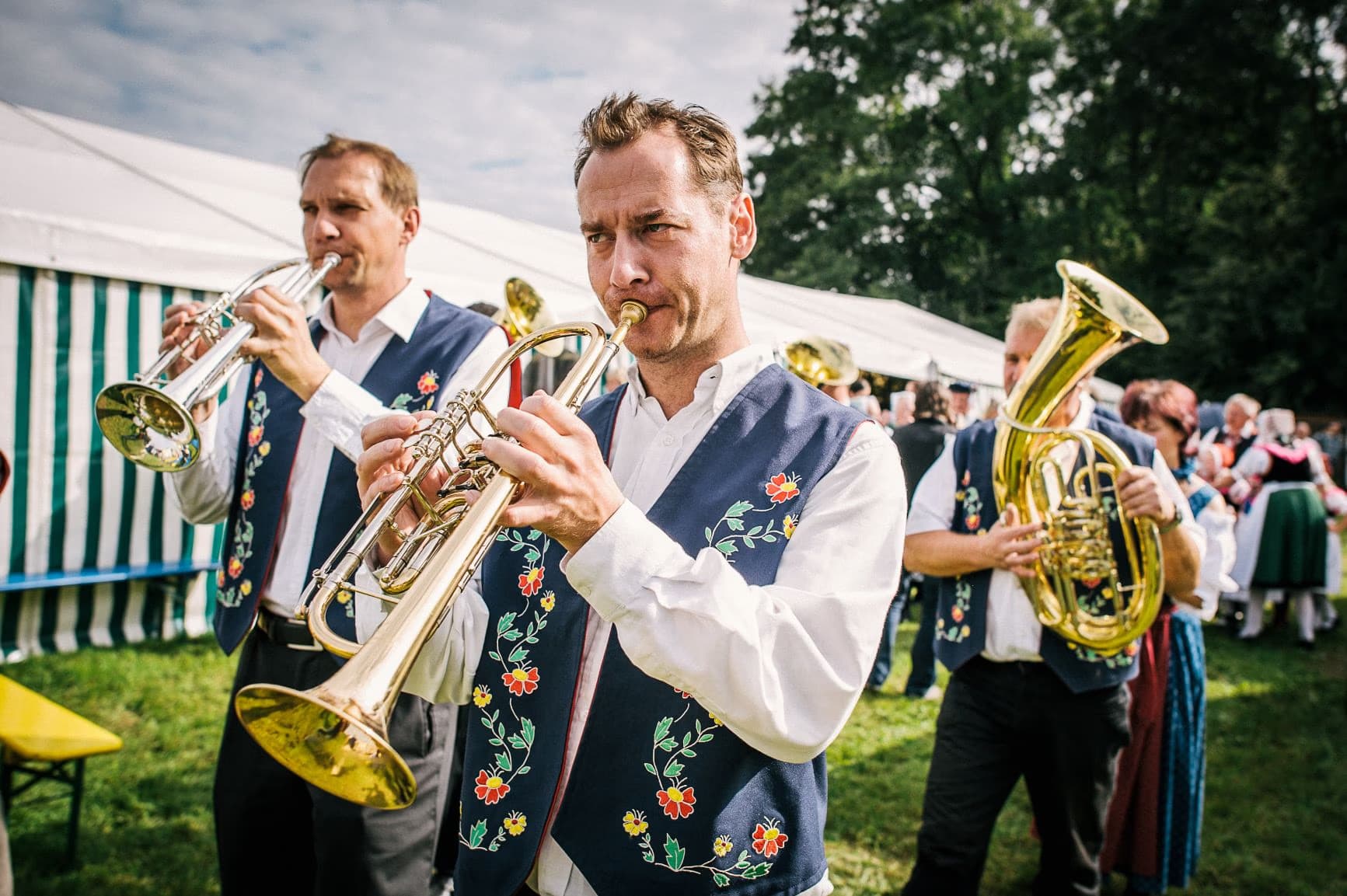 Musicians in traditional folk vests play brass instruments at an outdoor festival tent event.