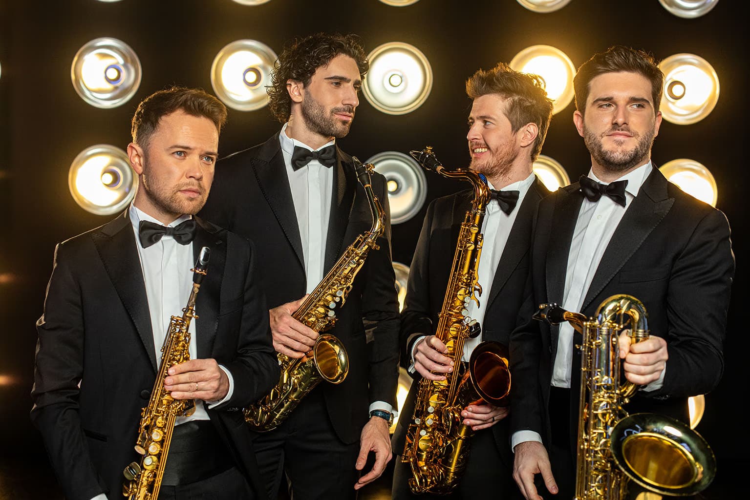 Four men in black tuxedos hold saxophones against a dark background with circular spotlights.