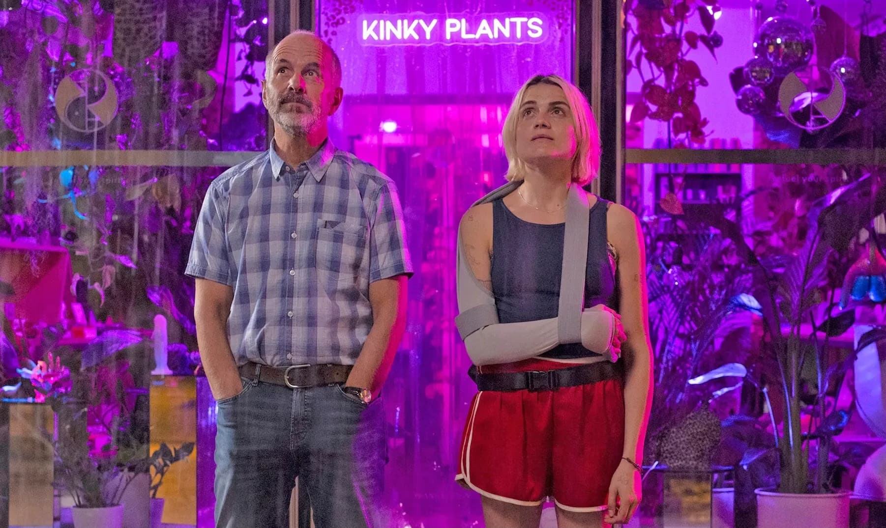 Ein Mann mittleren Alters und eine Frau mit verletztem Arm stehen vor einem Laden namens Kinky Plants.