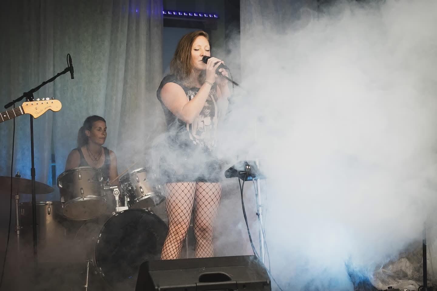 A female singer performs on stage with a drummer behind her surrounded by thick smoke.