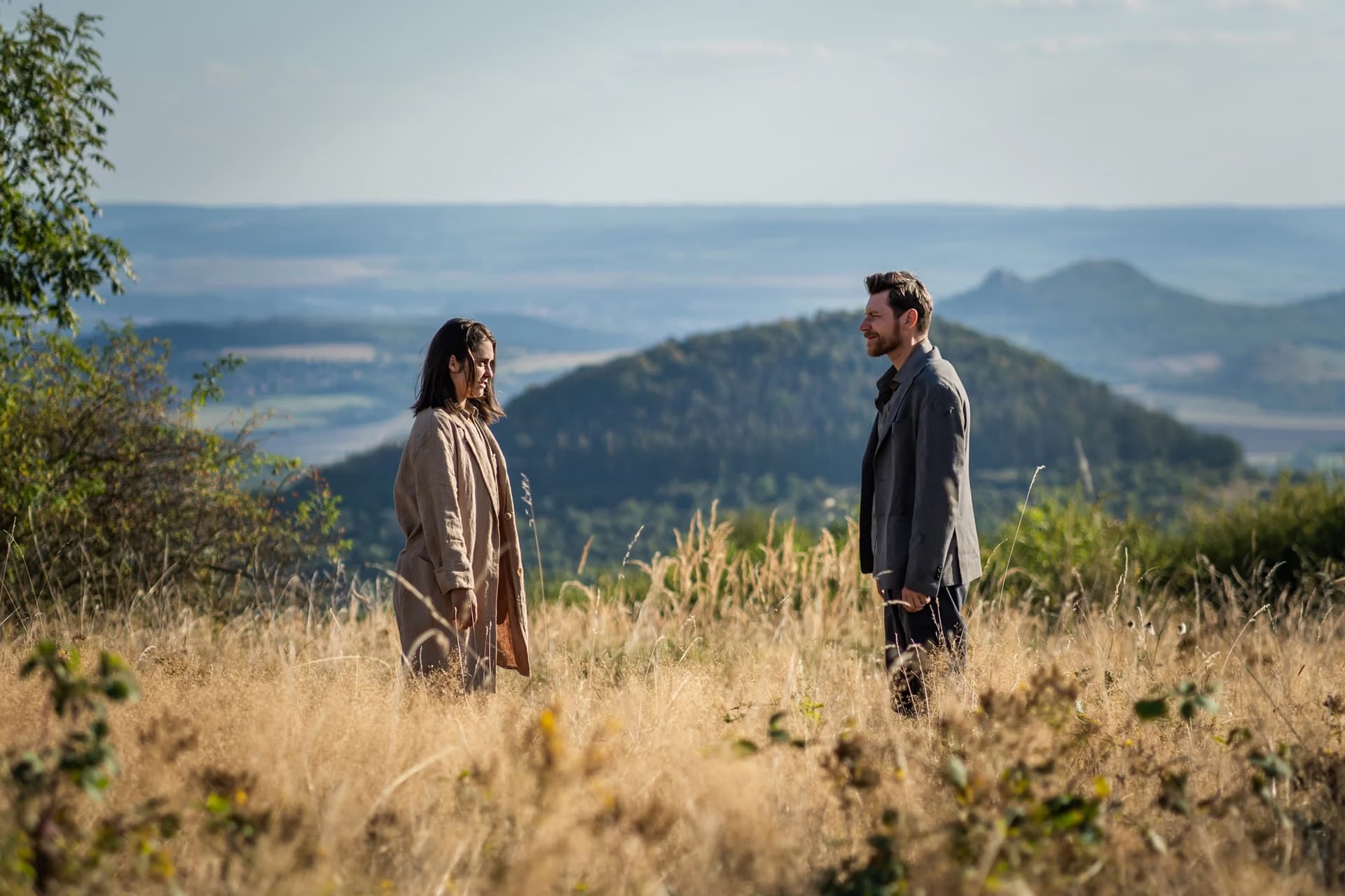 A man and woman stand facing each other in a grassy field with hills in the background.