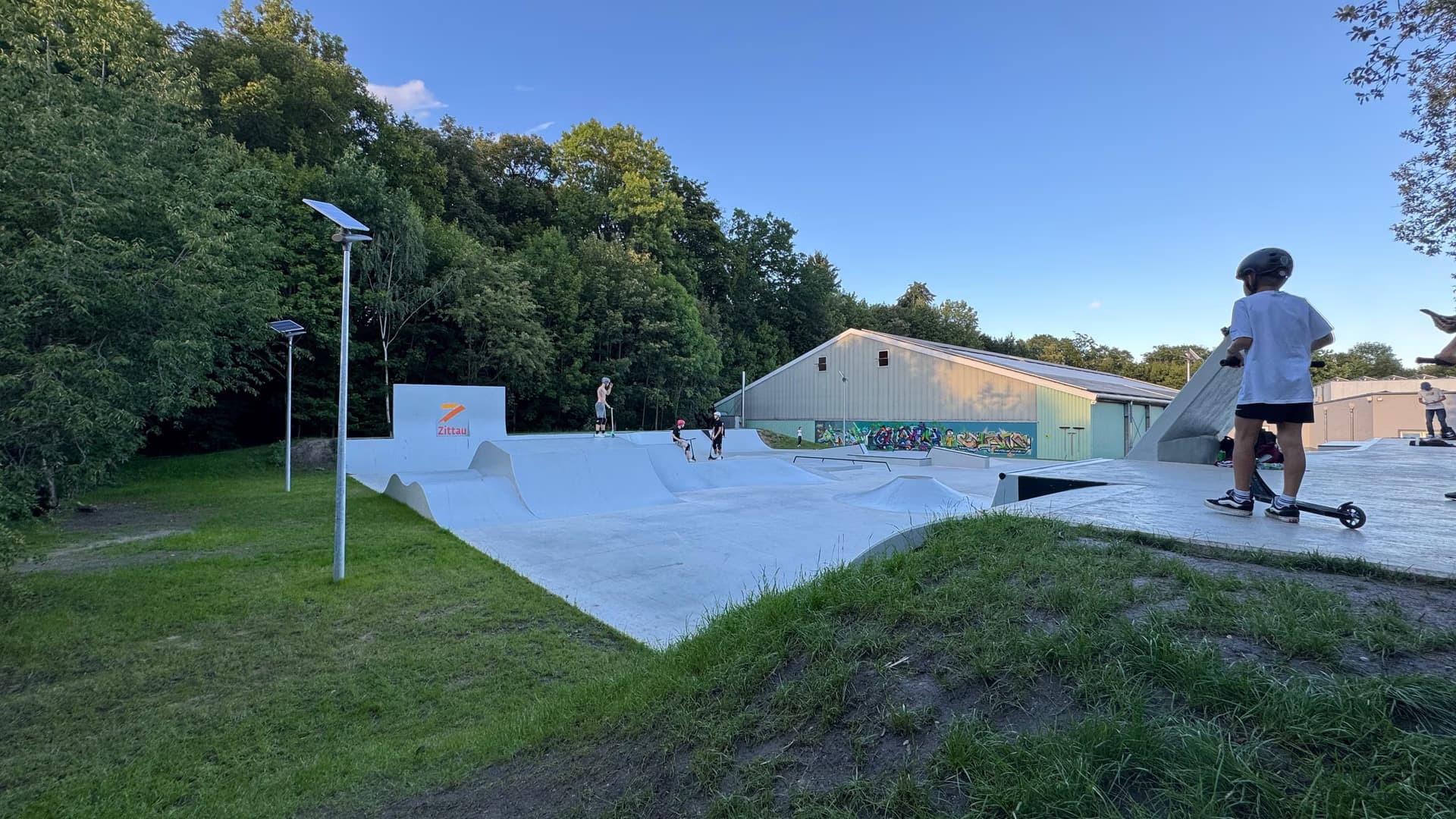 Ludzie jeżdżą na hulajnogach po betonowym skateparku obok trawiastego zbocza i drzew.
