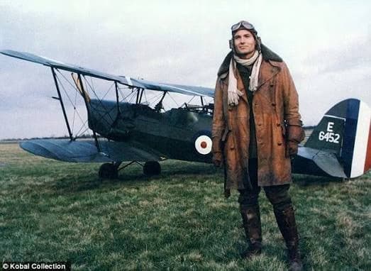 A pilot in leather coat and flight goggles stands before a vintage biplane on a field.