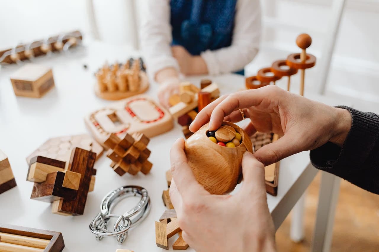 Hands are assembling various wooden mechanical brain teaser puzzles on a white table.