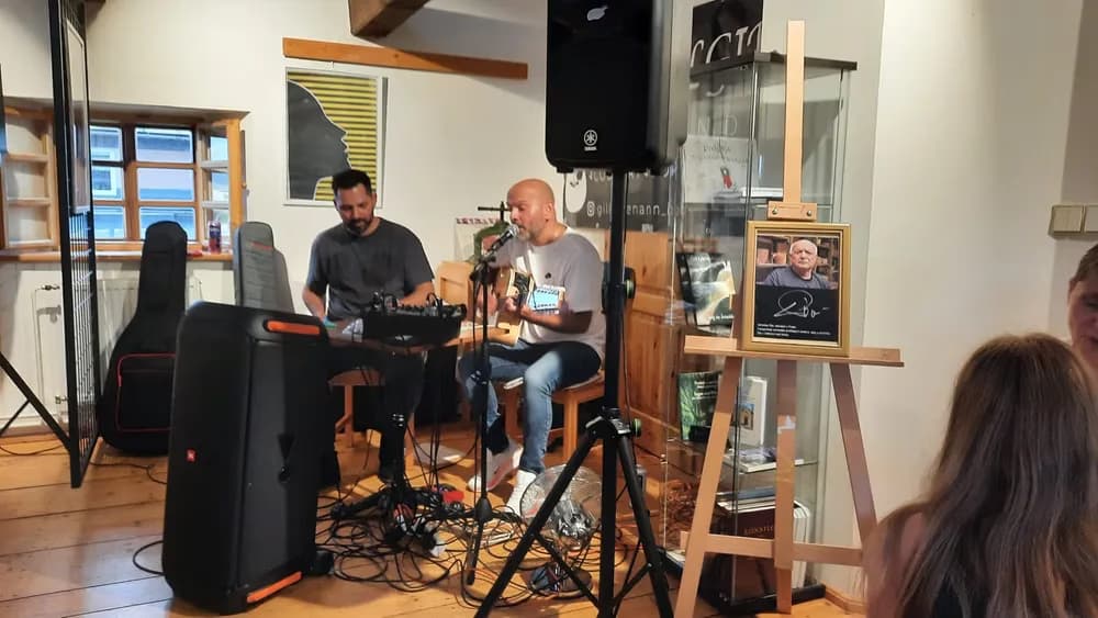 Zwei Männer spielen in einem Raum akustische Musik, daneben steht eine Staffelei mit einem gerahmten Porträt.