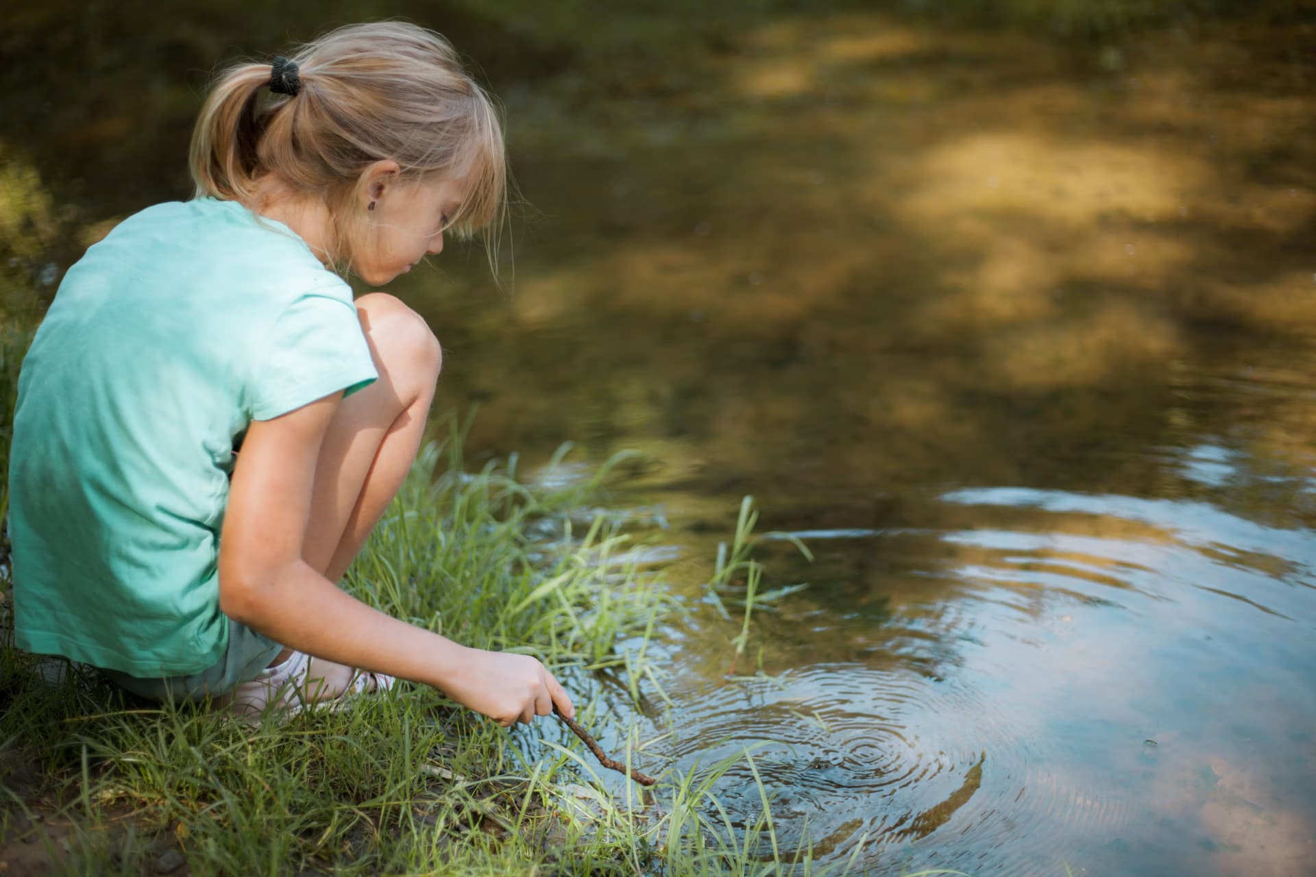 Young girl in a light blue shirt playing with a stick in a calm river.