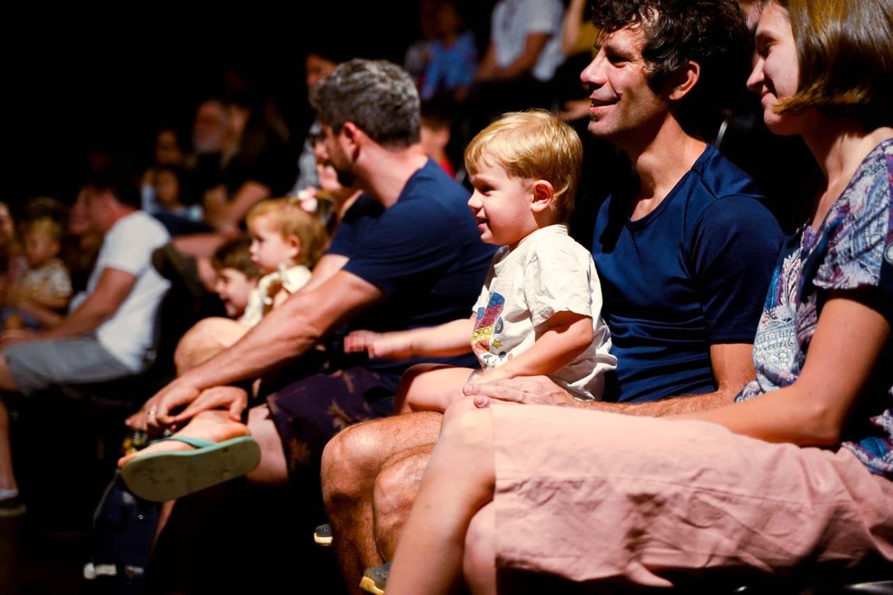 A group of families sits in a dimly lit theater watching a performance.