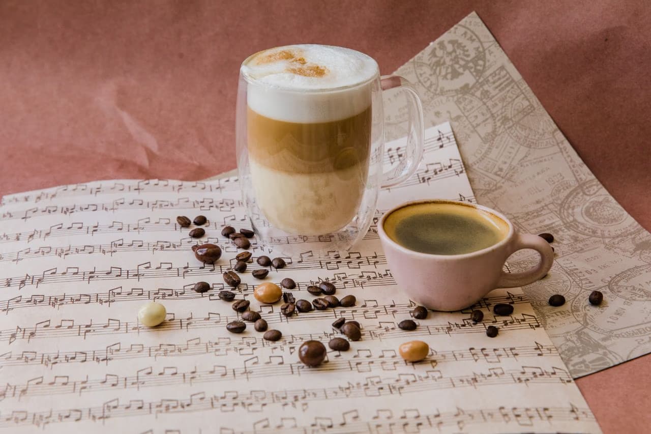 A glass latte and a small black coffee sit on a sheet of music with scattered coffee beans.