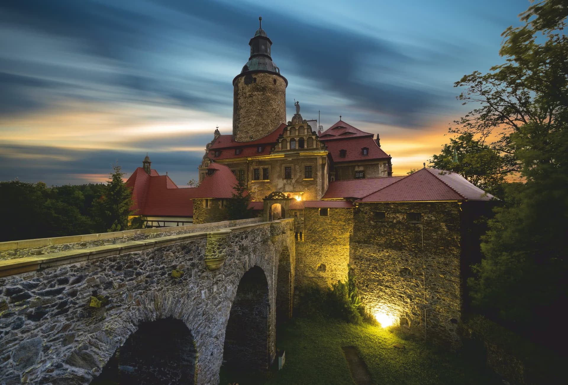 A stone castle with a tower and bridge illuminated against a dramatic sunset sky.