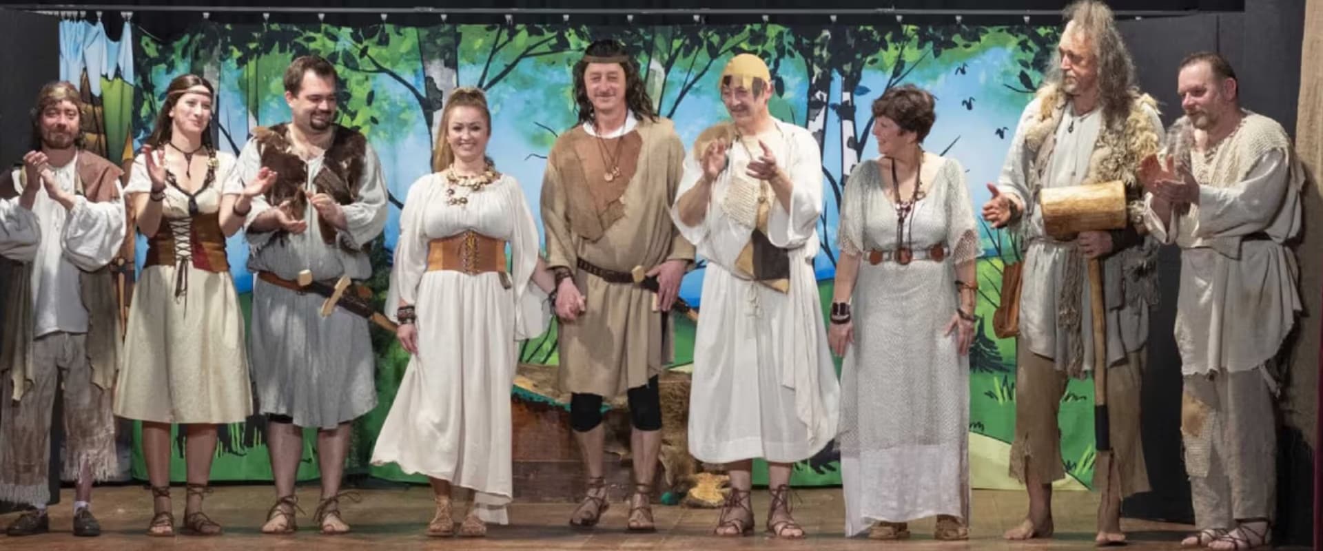 Actors in historical costumes stand in a row on stage in front of a painted forest backdrop.