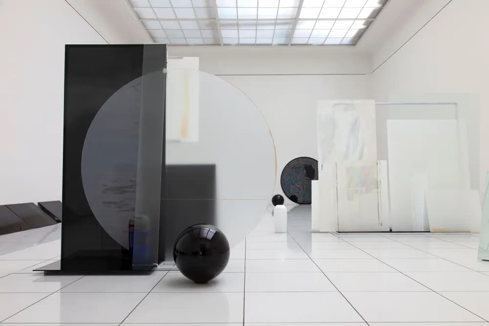 Abstraktní umělecká instalace s černými geometrickými tvary na bílé dlažbě v galerii.