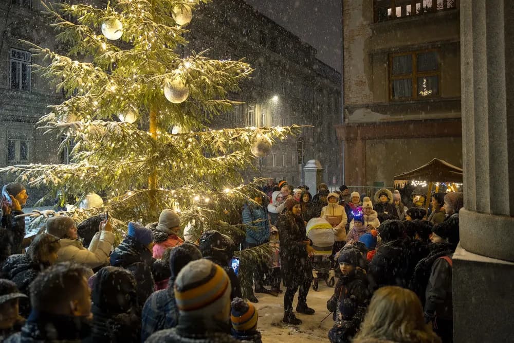 A crowd gathers around a brightly lit Christmas tree in a snowy town square at night.