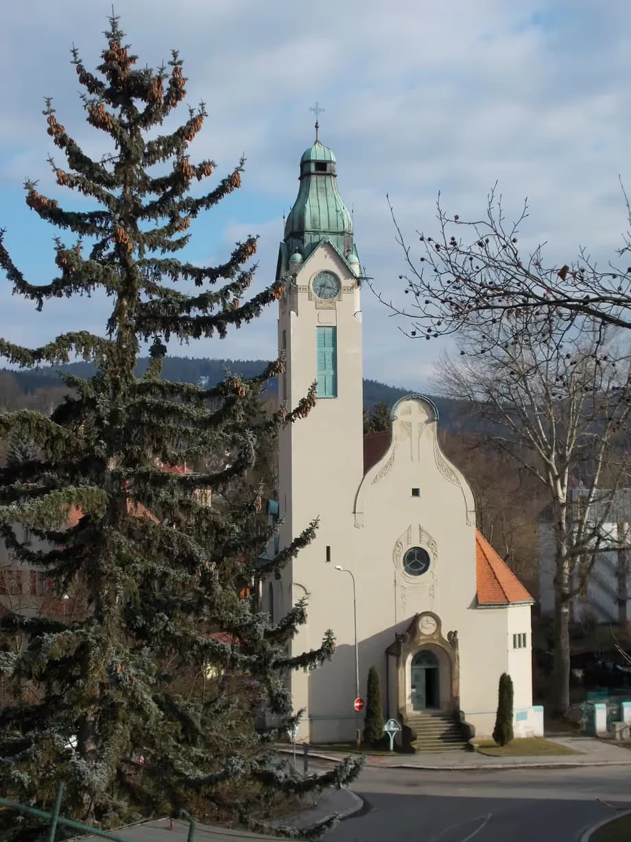 Kostel s vysokou věží stojí nápadně u vzrostlého jehličnatého stromu.