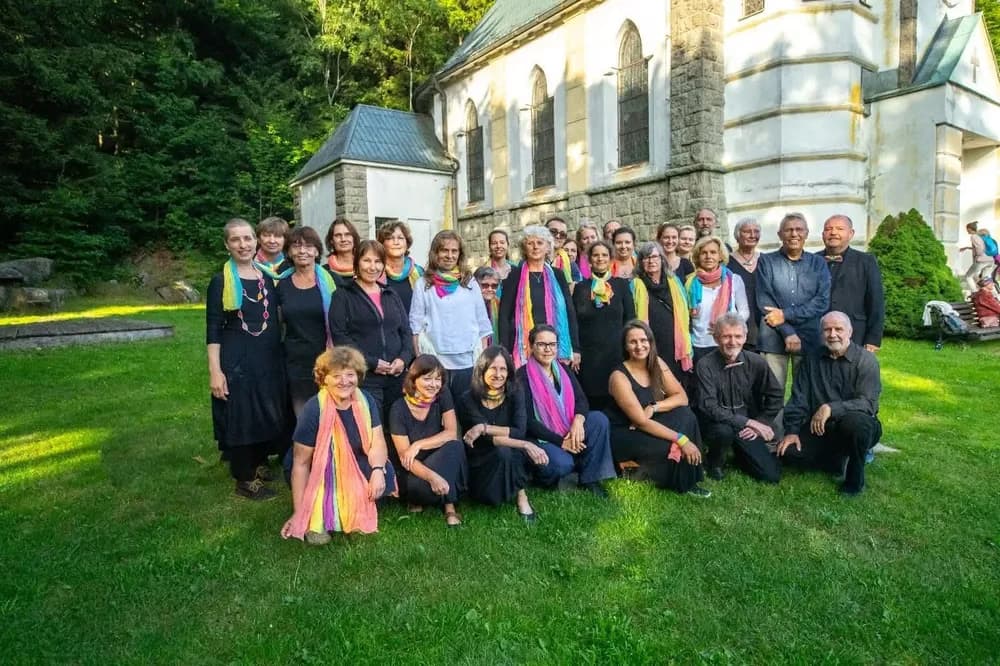 A large choir group posing in front of a stone church on a grassy lawn.