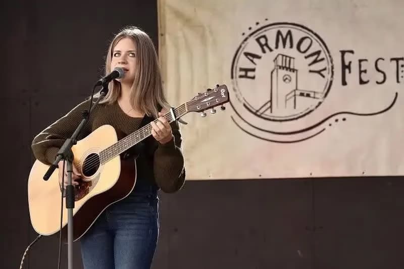 Young woman playing acoustic guitar and singing on stage in front of a Harmony Fest banner.