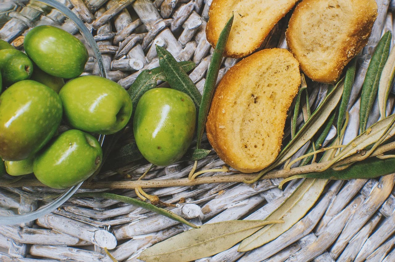 Green olives in a glass bowl sit next to bread slices and olive branches on a woven surface.