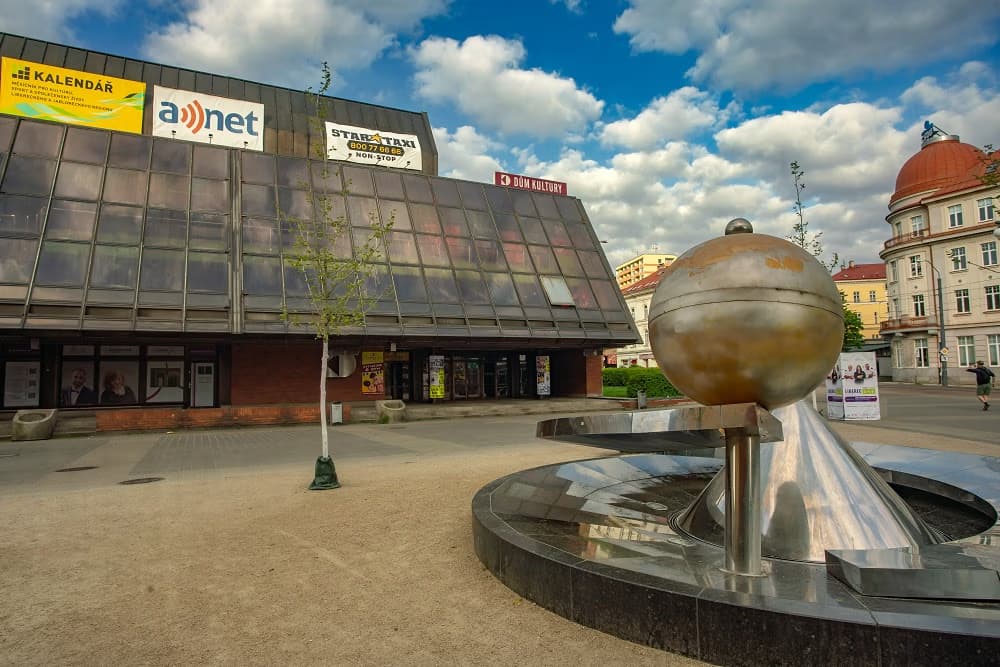 A modern building with glass panels and a spherical fountain in a town square under blue skies.