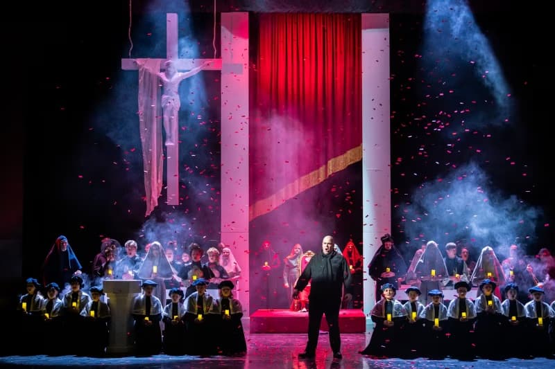 A man stands center stage during a theatrical performance with actors in robes holding candles.