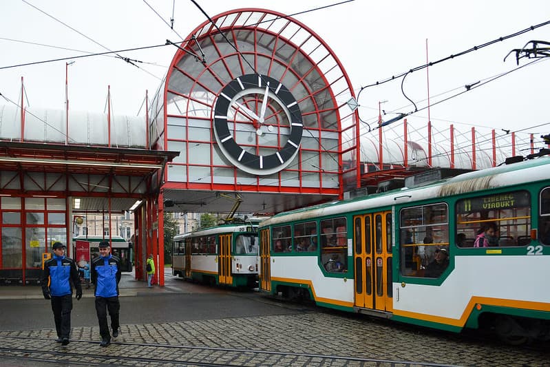 Two uniformed men walk in front of a tram terminal featuring a large clock.
