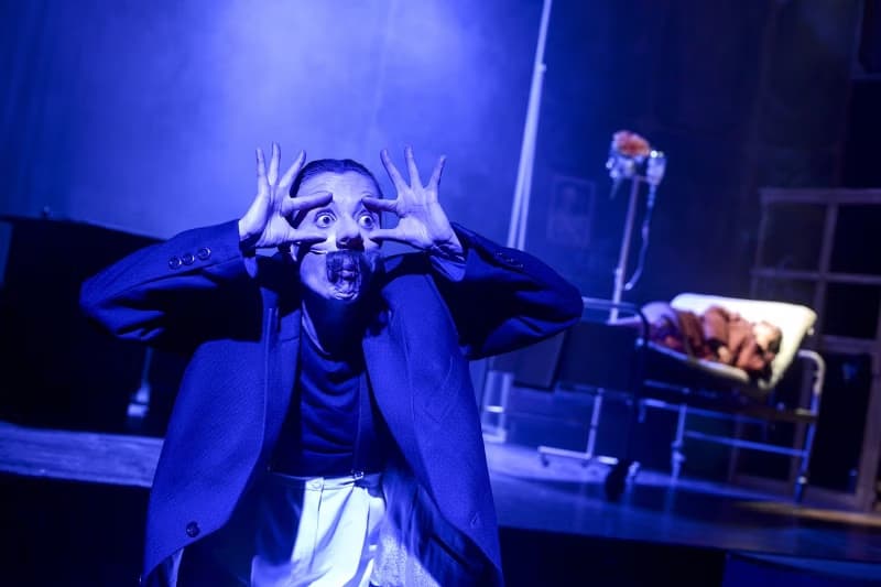 An actor in a dark suit makes a surprised gesture on a dimly lit theater stage.