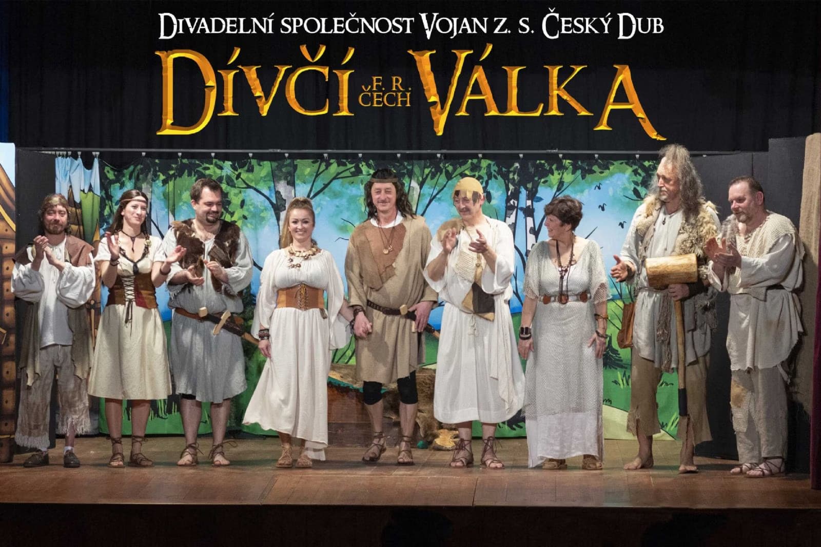 Theater troupe in historical costumes stands on stage before a painted forest backdrop.