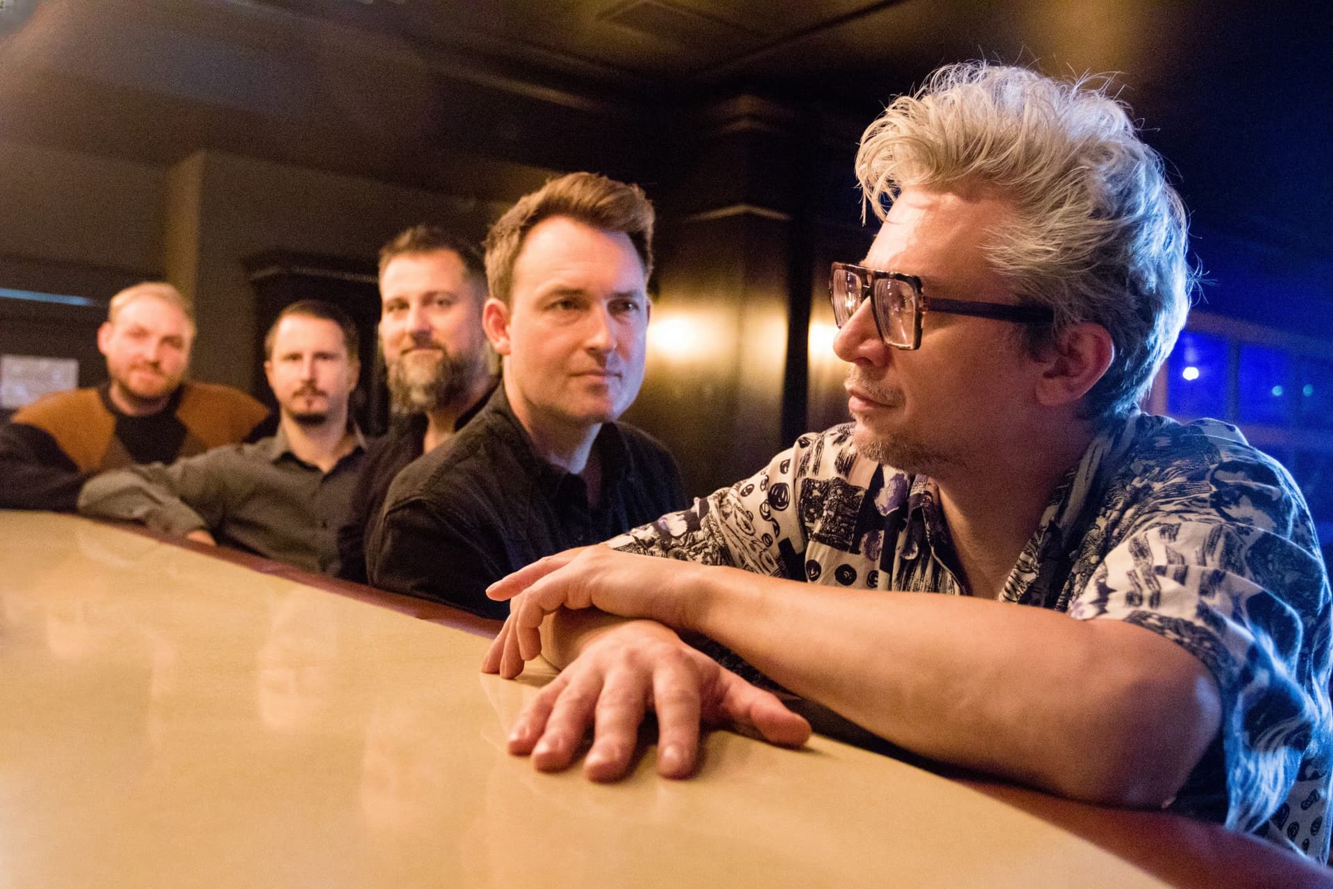 Pět mužů v ležérním oblečení stojí a opírá se o dřevěný bar v šeru hospody.