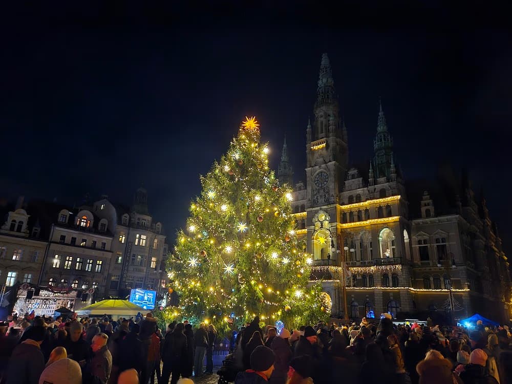 A large illuminated Christmas tree stands in a town square before a historic building at night.