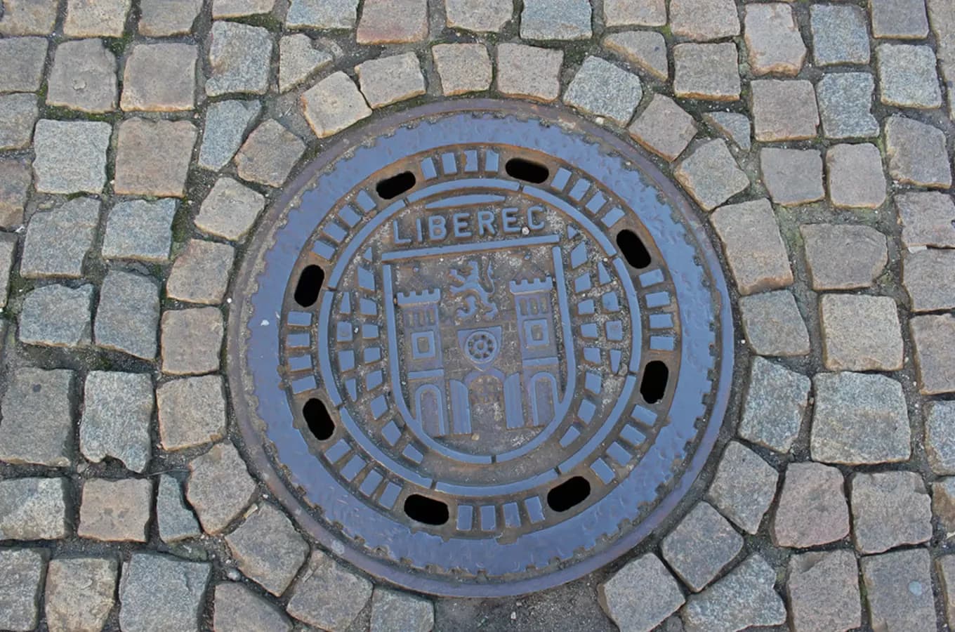 A metal manhole cover with the Liberec coat of arms set in a cobblestone street.
