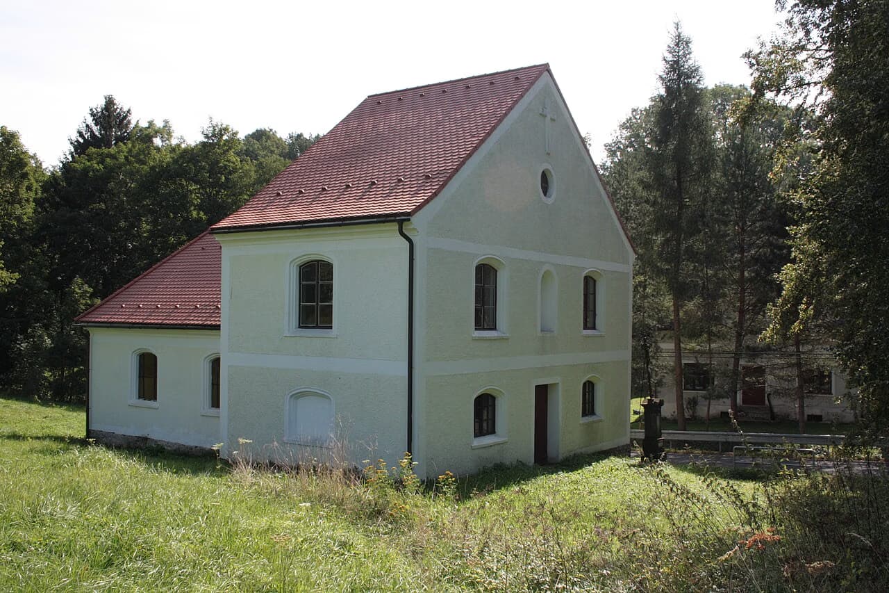 A historic white building with a red tiled roof stands in a grassy field surrounded by trees.