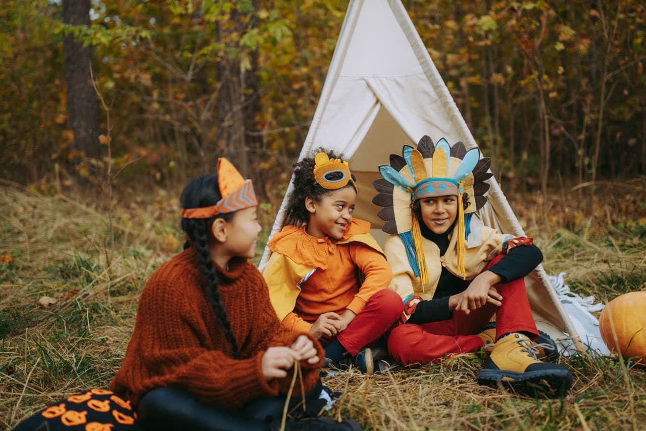 Three children in various costumes sit on the grass next to a small white tent.