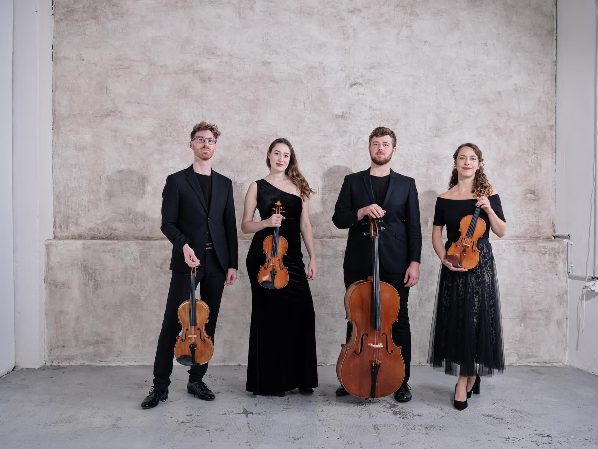 Four classical musicians in elegant black formal wear pose with violins and a cello against a concrete wall.