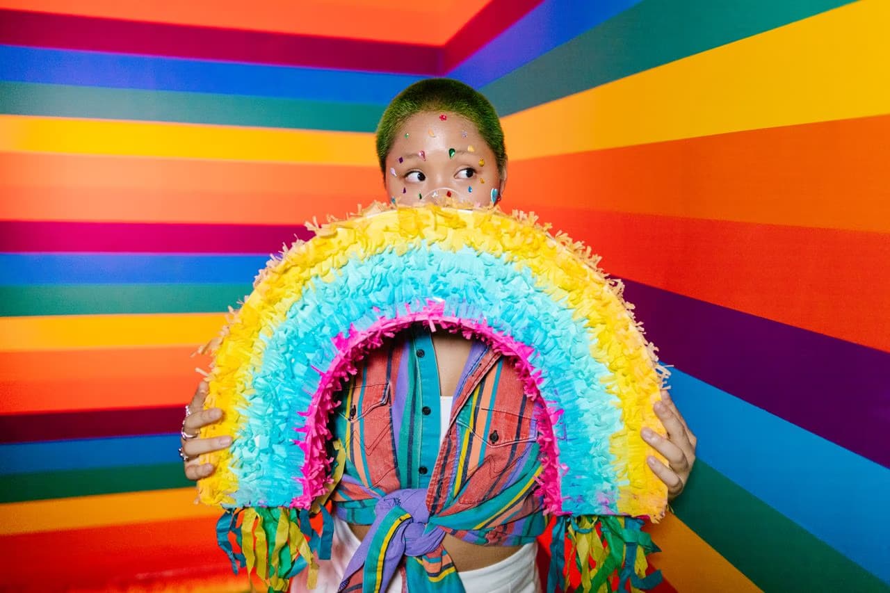Person with green hair and face glitter holds a colorful rainbow piñata against a striped background.