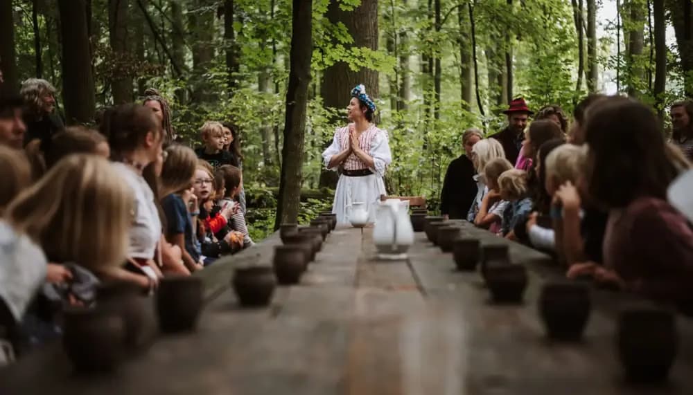 Osoba w białym stroju stoi na czele długiego stołu wśród ludzi w lesie.