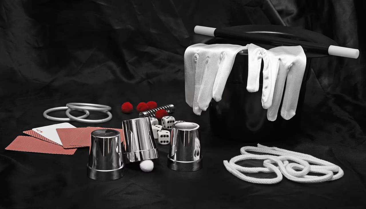 Magic props including a top hat, white gloves, metal cups, dice, cards, and rope on black.