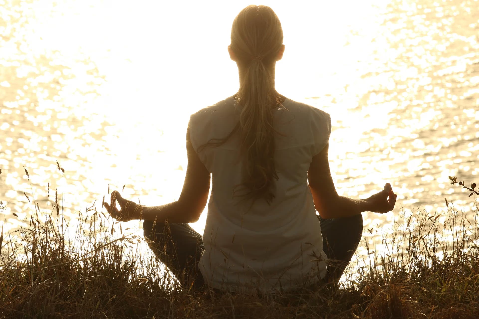 A woman sits in a meditative pose on a riverbank during a golden sunset.
