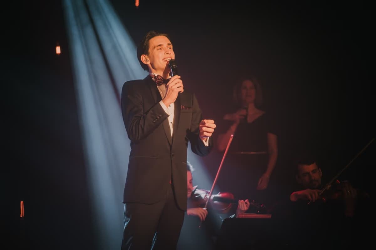 A man in a tuxedo sings into a microphone on stage, illuminated by a spotlight.