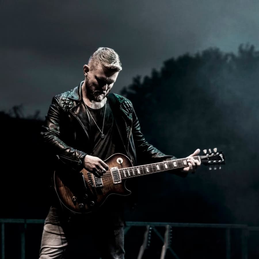 Mężczyzna z krótką brodą gra w nocy na scenie na brązowej gitarze elektrycznej.