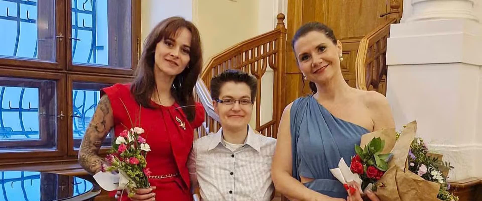 Three people holding flowers pose for a photo in an indoor setting with wooden railing.