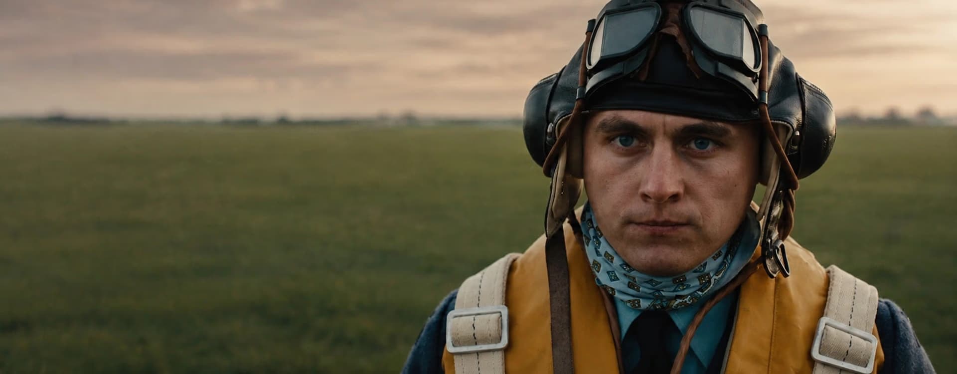 Stíhací pilot v historické letecké výstroji stojí na travnatém poli při západu slunce.