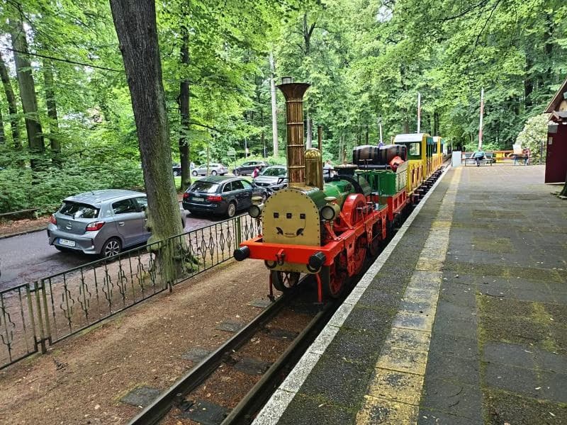 A small colorful passenger train sits on tracks at a forested station platform.